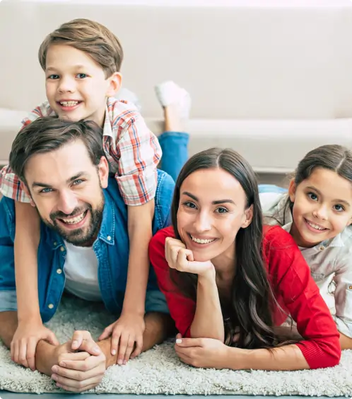 A smiling family of four lies on a carpet in a living room. The father and mother are in front, with their son lying on the father’s back and their daughter next to the mother. All are looking at the camera happily.