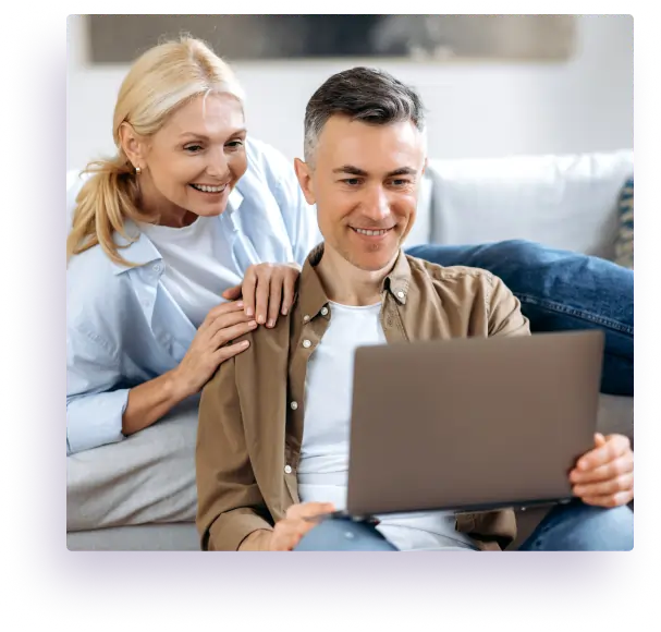 A smiling middle-aged couple sits on a couch at home, looking at a laptop screen together. The woman stands behind the man, resting her hand on his shoulder, both appearing engaged and happy.